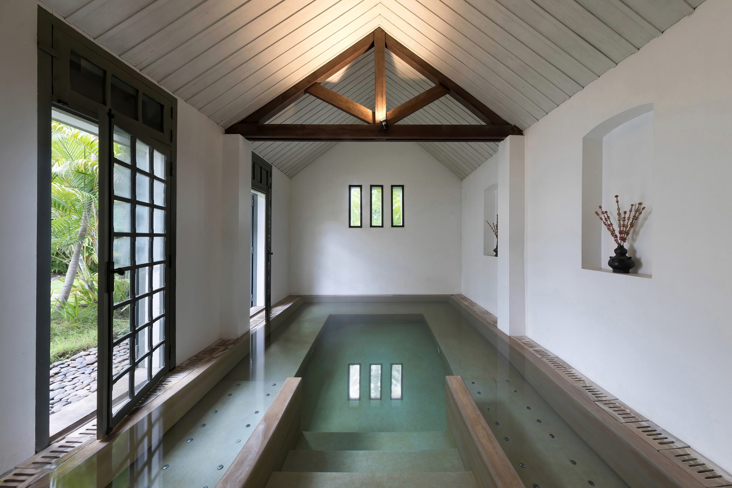 Indoor spa pool in a white vaulted room, timber doors open to planting outside